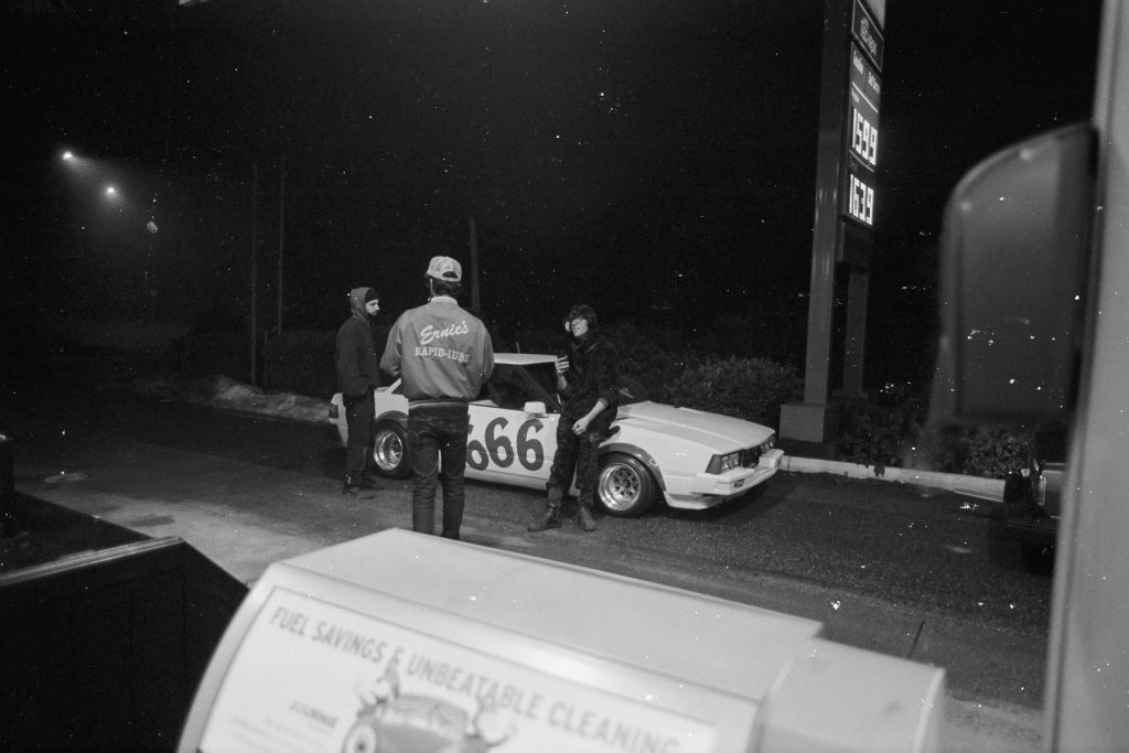 Three men gather around a vintage car with the number 666 by gas prices at a night-time gas station. Sign reads Ernie's Rapid-Lube.