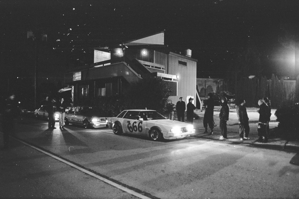 Night street scene with parked classic cars and people standing nearby, including a car labeled 666 out front.