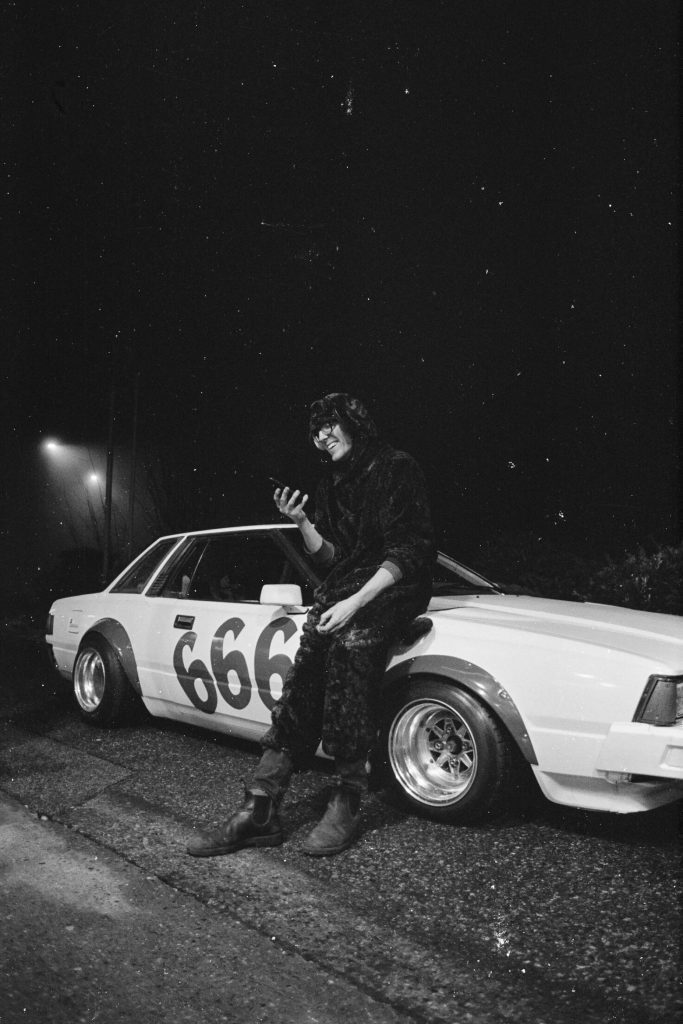 Man in a hooded outfit sitting on a white sports car with 666 painted on the side, smiling and using his phone.