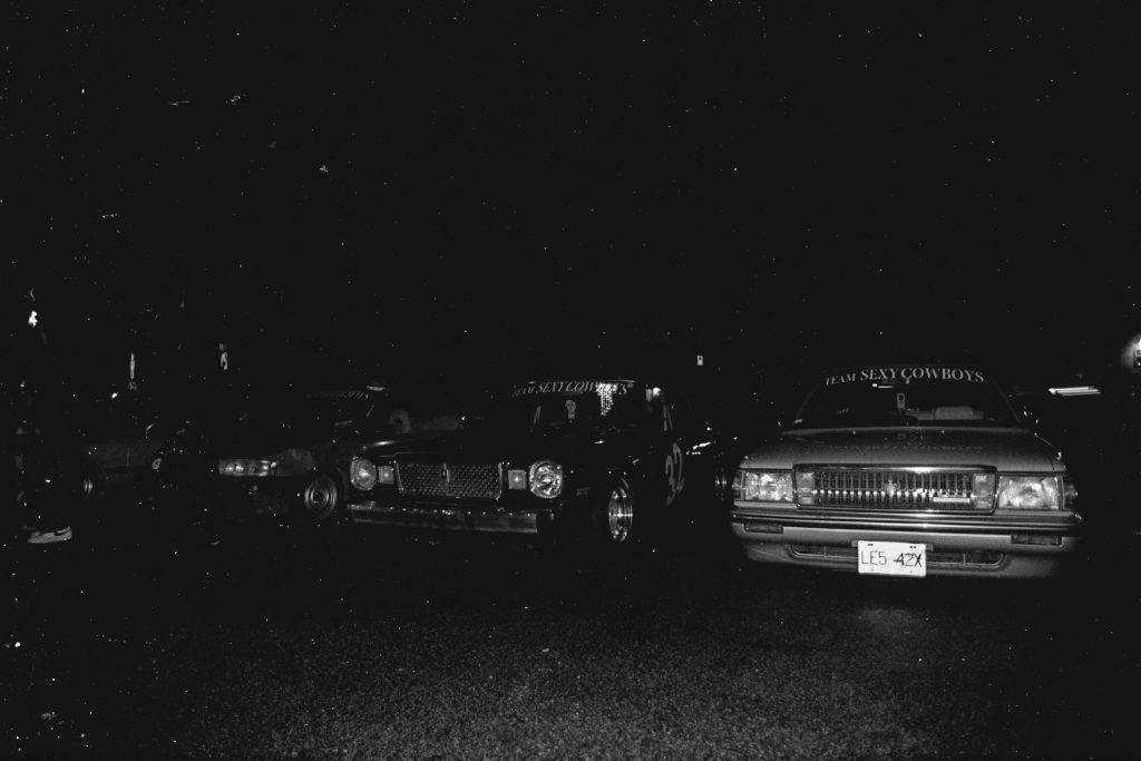 Vintage cars at night displaying Team Sexy Cowboys banners on windshields, showcasing car culture.