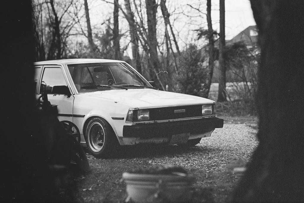 Vintage white Japanese car parked outdoors in a wooded area, showcasing classic design and scenery.