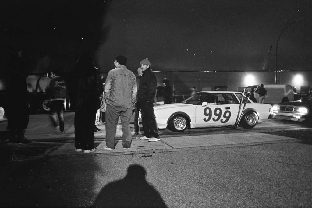 Street scene at night with people around a car numbered 999, under streetlights, one worn jacket reads スピードパンサー (Speed Panther).