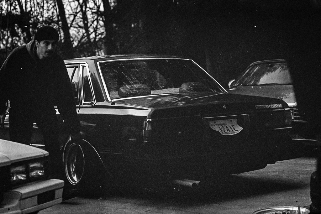 A man standing next to a classic car in a dark, wooded area with a Washington license plate, partially visible.
