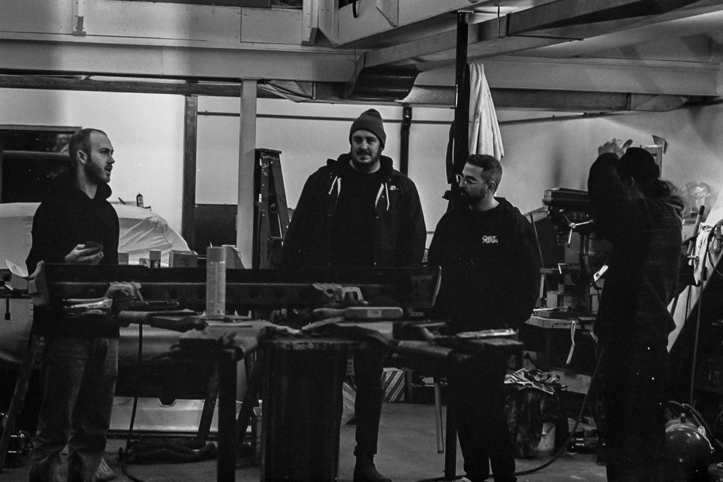Four men in a workshop discussing a project with tools and equipment around them, monochrome image.
