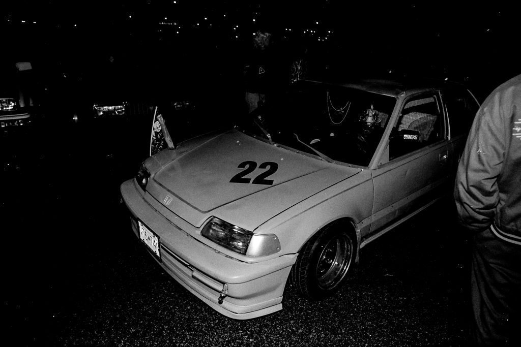Black and white image of a modified Honda car with the number 22, parked in a night setting with people around.