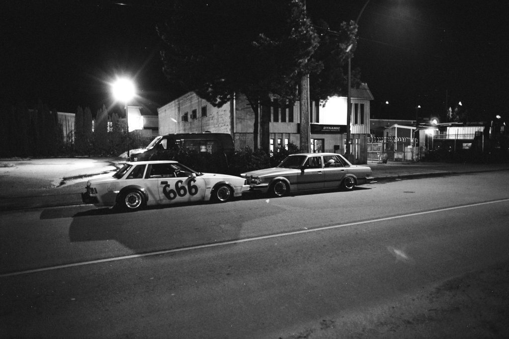 Two vintage cars parked at night in front of a building labeled DYNAMIC; one car has 666 painted on its door.