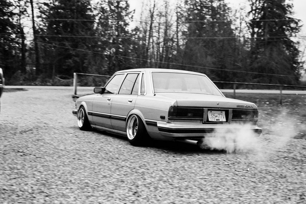 Black and white image of a vintage Toyota sedan drifting on a gravel road with smoke arising from the tires.