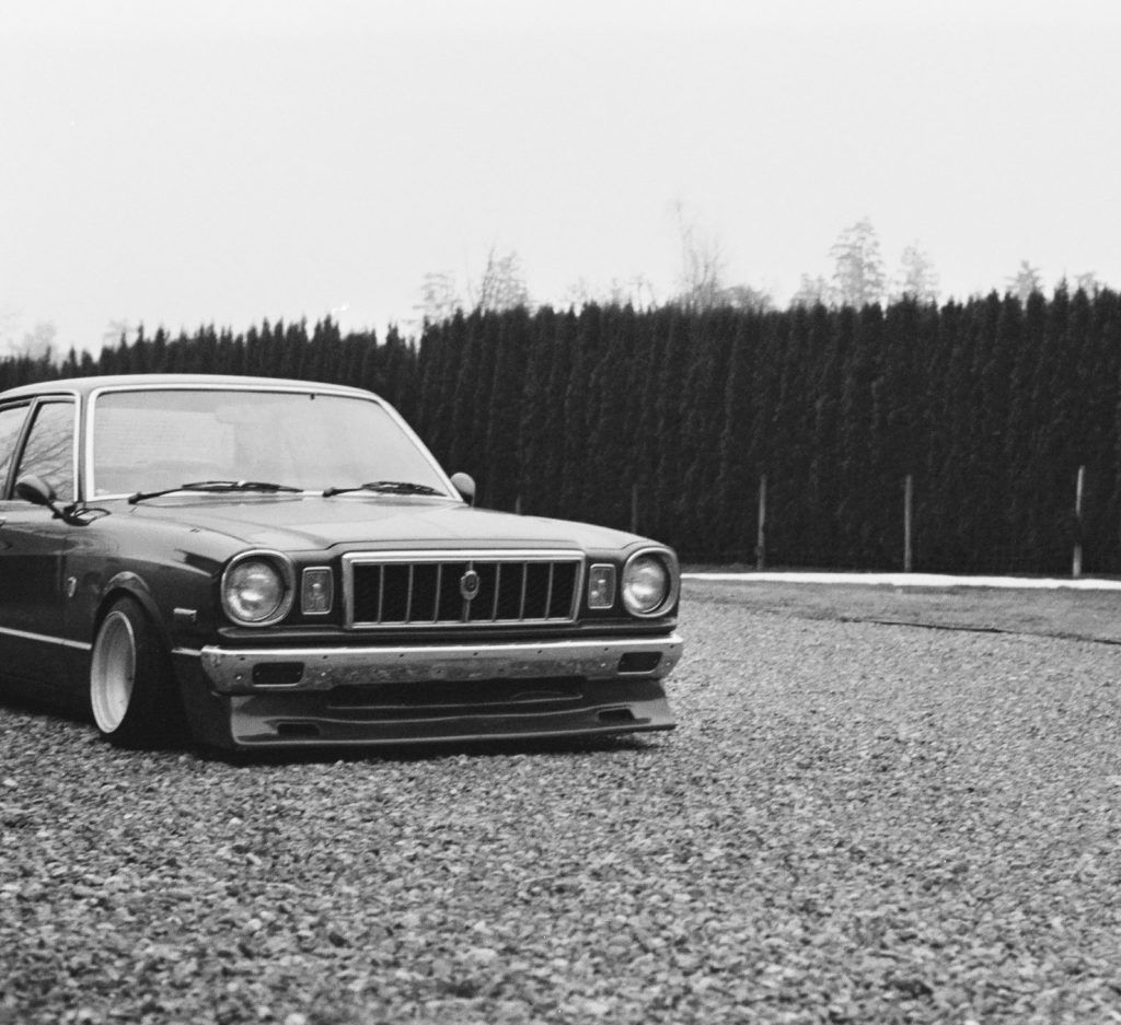 Monochrome image of a vintage, lowrider Japanese car parked on gravel, flanked by tall hedge plants in the background.