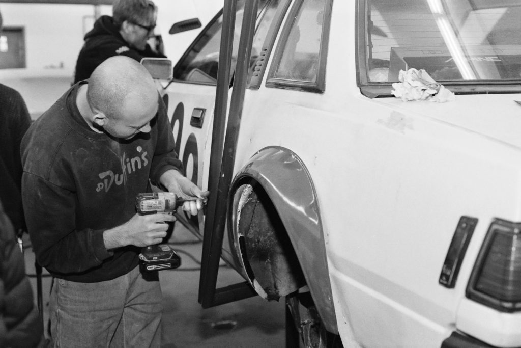 Mechanic working on a race car with a power drill in a garage. Person in background blurred. Number 20 on car door.