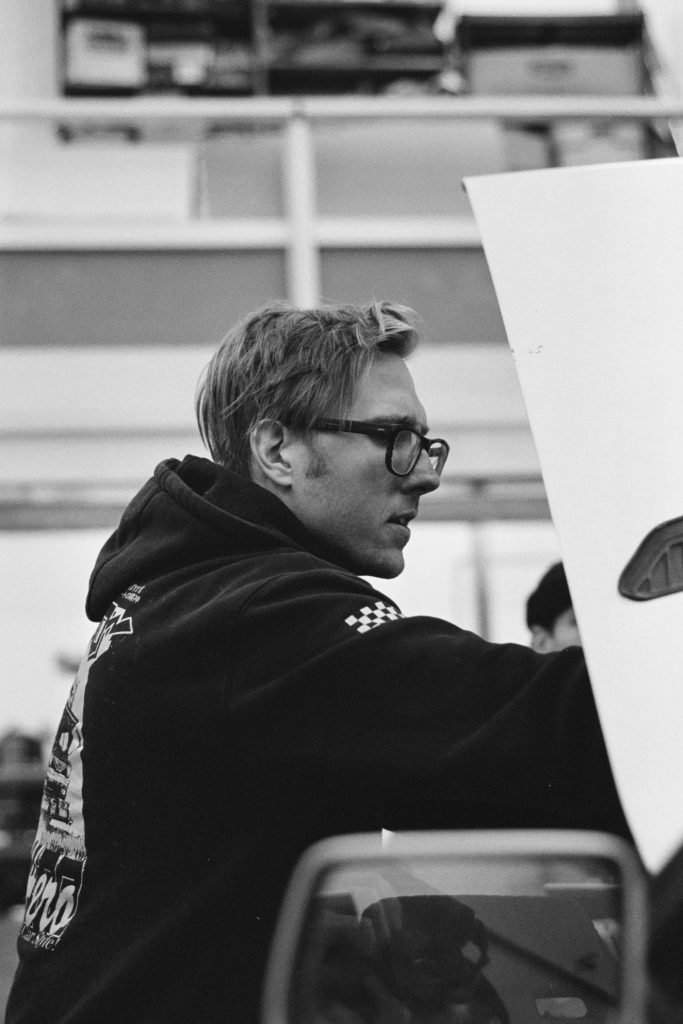 A person working under a car hood in a garage, wearing glasses and a hoodie with Japanese text オート (Auto).