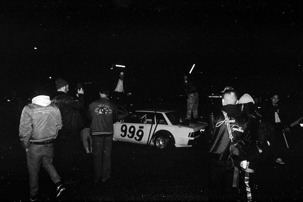 Car enthusiasts and a vintage race car #999 at a night car meet. One jacket reads Alone Together in Japanese translation.