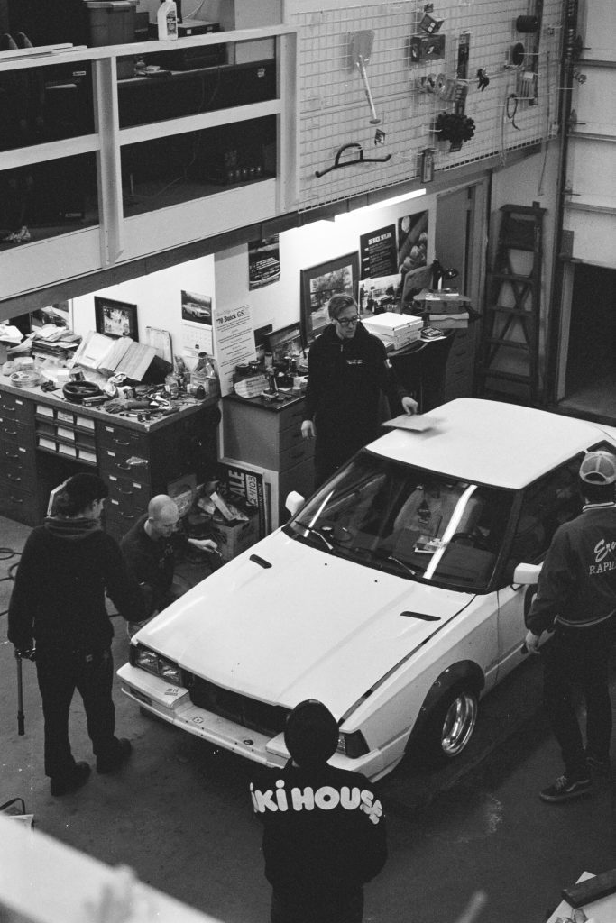 Group of mechanics working on a white car in a garage with iKi HOUSE text on one jacket.