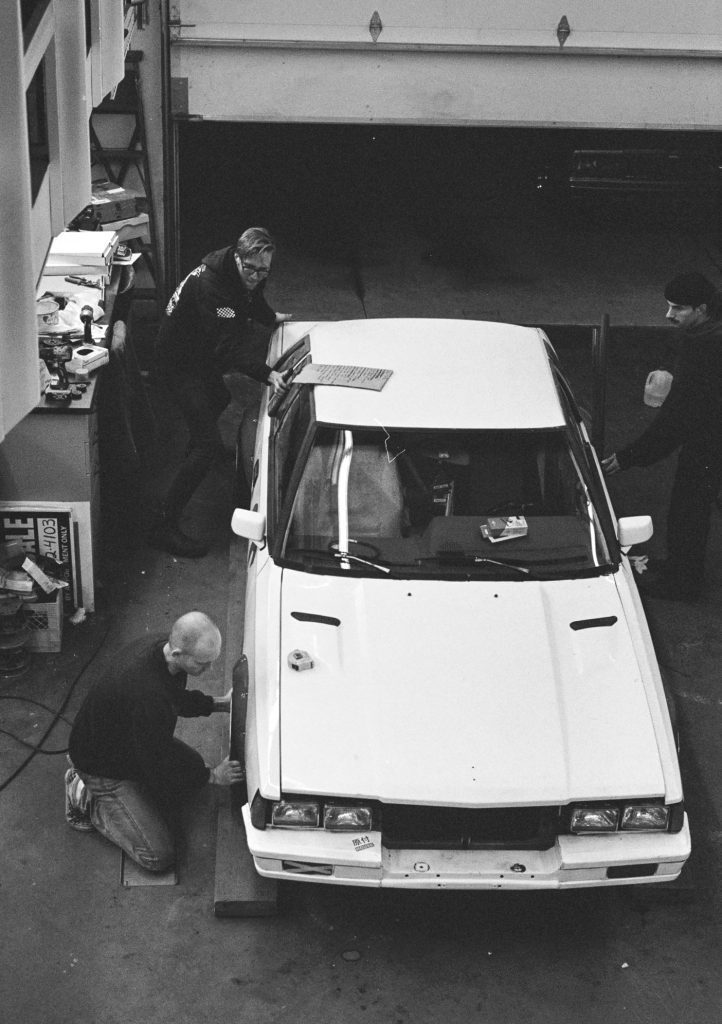 Mechanics working on a white car in a garage, with a Japanese Inspection label on the front bumper.