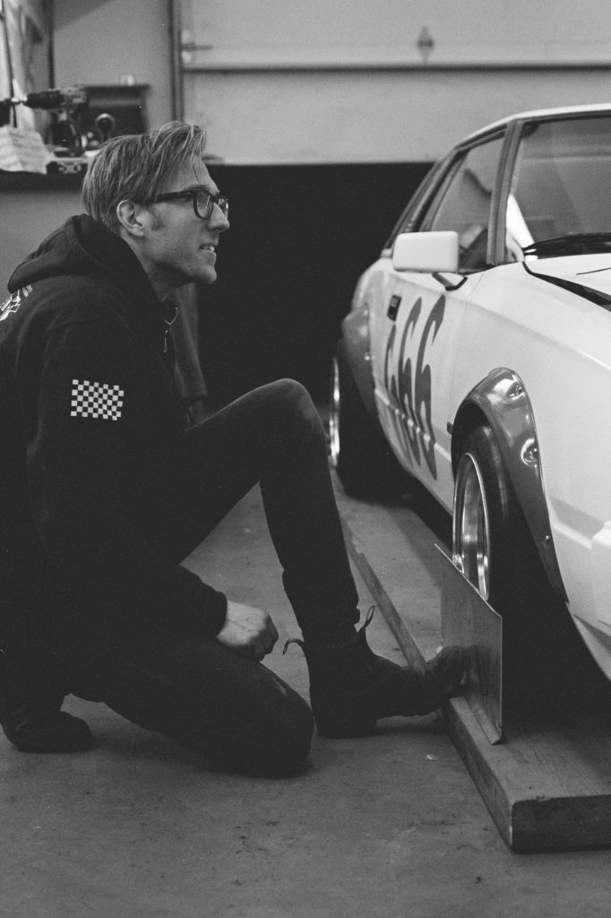 Man adjusting a racing car in a garage, wearing glasses and a black hoodie with a checkered patch on the sleeve.