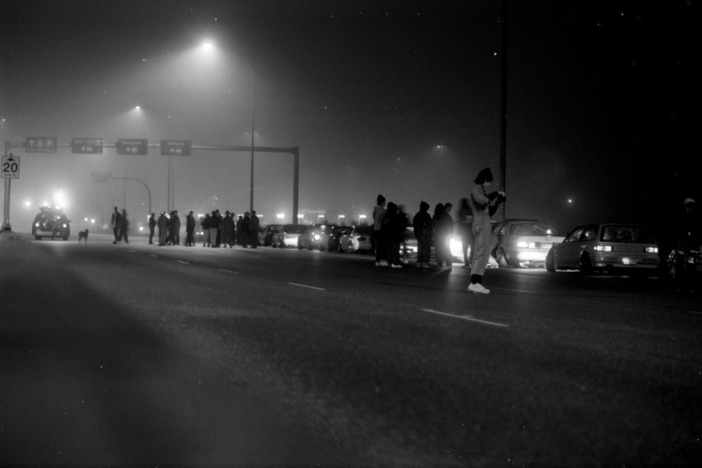 A late-night gathering of people and cars under streetlights in a foggy environment, with signs and 20km speed limit visible.