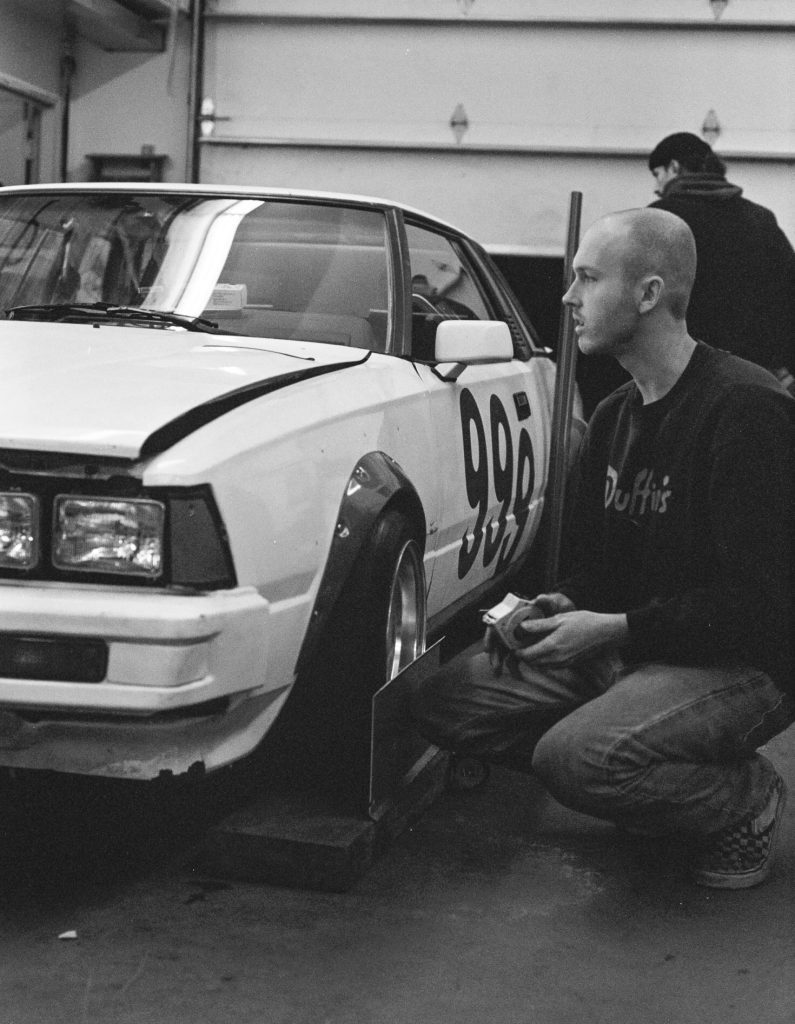 Man inspecting a race car numbered 996 in a garage, wearing a Duffis shirt, while another person works in the background.