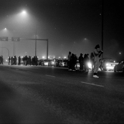 A late-night gathering of people and cars under streetlights in a foggy environment, with signs and 20km speed limit visible.