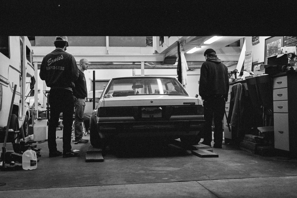 Three mechanics working in a garage, inspecting a vintage car with Ernie's Rapid-Lube on a jacket. Black and white photo.