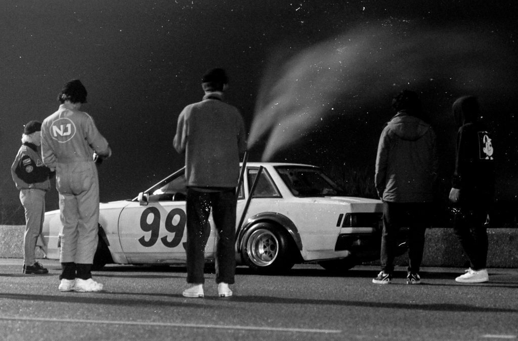 Vintage racing car number 99 with a group of people in racing gear at night, steam emitting from the car's rear.