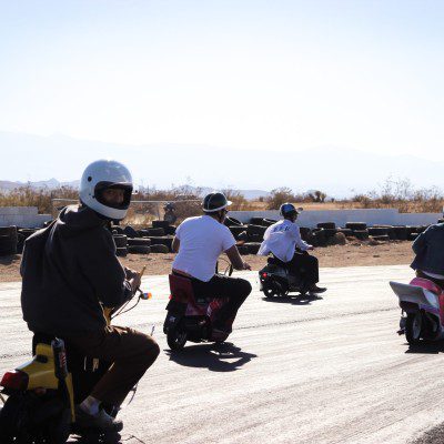 Group of people in helmets riding scooters on a sunny day, with desert landscape and tires in the background.