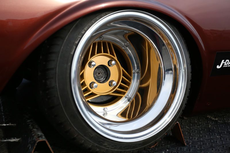 Close-up of a custom car wheel with gold spokes and polished silver rim next to JDM sticker on a brown vehicle.