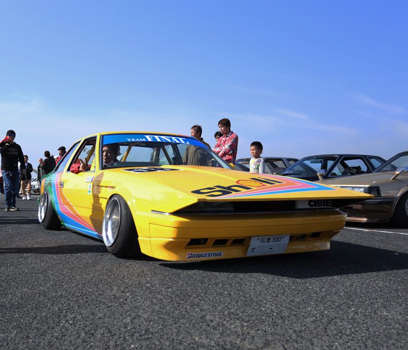 Yellow custom sports car with multicolor stripes at an outdoor car show in Japan, with spectators in the background.