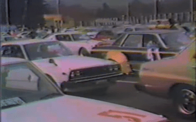 Vintage Japanese car meetup in a parking lot with various classic vehicles and scenery in the background.