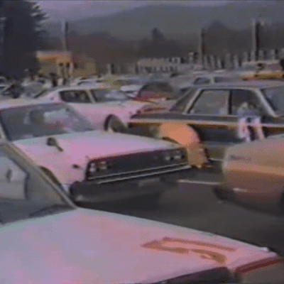 Vintage Japanese car meetup in a parking lot with various classic vehicles and scenery in the background.