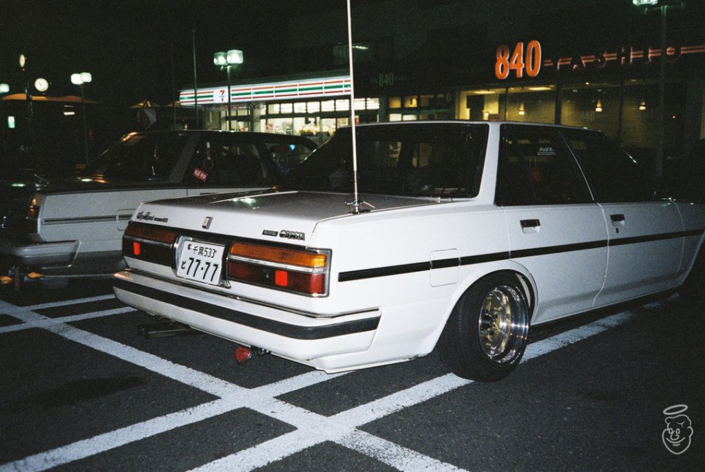 A white Toyota Chaser in a parking lot at night, near a 7-Eleven and next to a building with 840 on its sign.