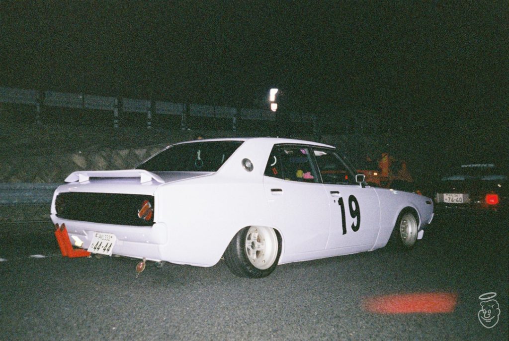 Classic white Japanese car with number 19 on the side parked at night, showcasing retro automotive style.