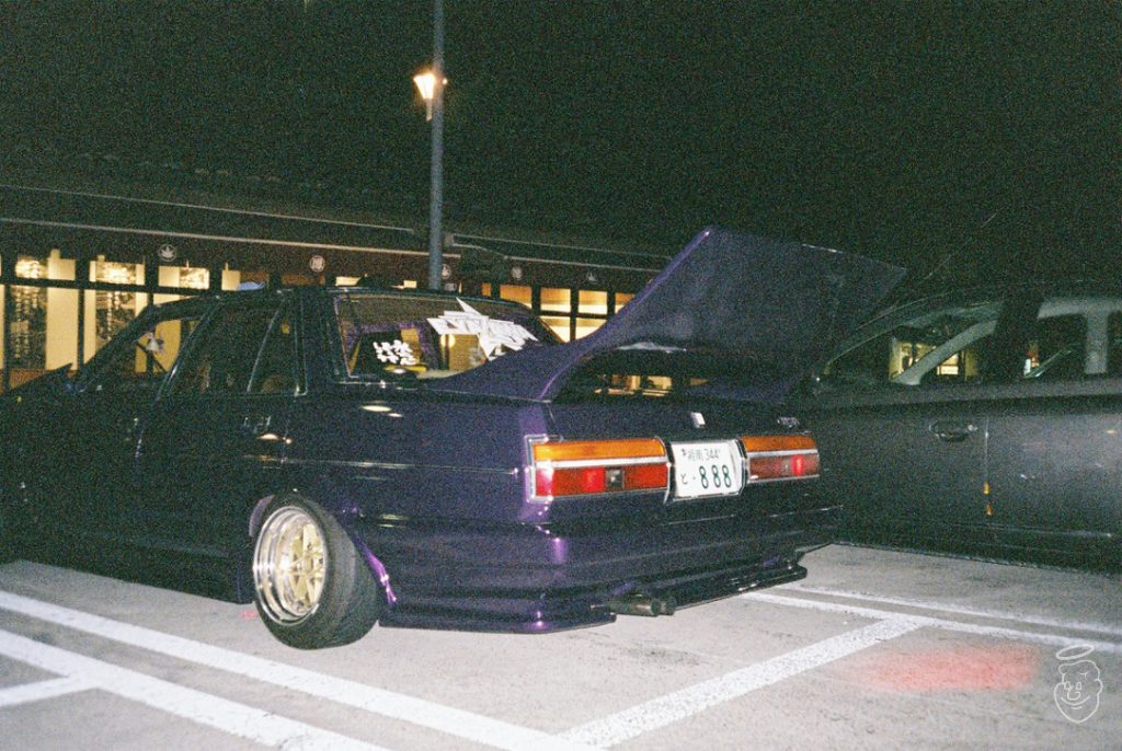Modified purple car with raised trunk, gold rims, and Japanese license plate, parked at night near a building.