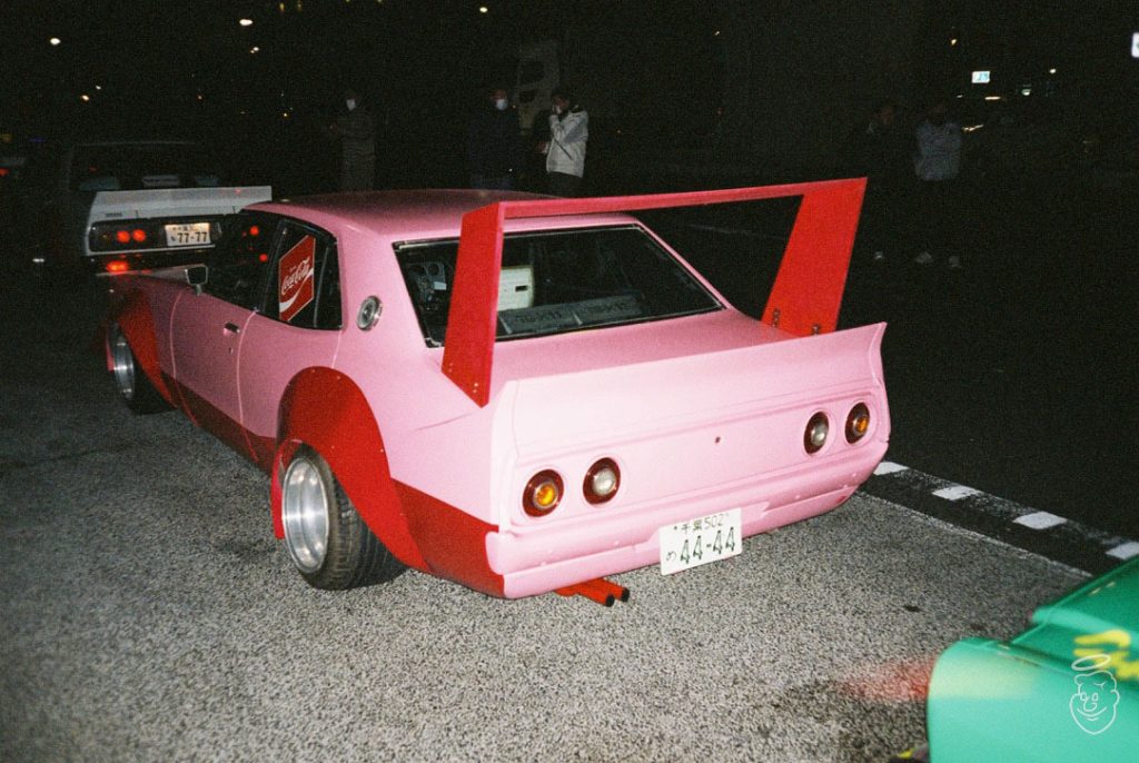 Custom pink and red Japanese Bosozoku car with large rear spoiler, in nighttime car meet. License plate reads 44-44.