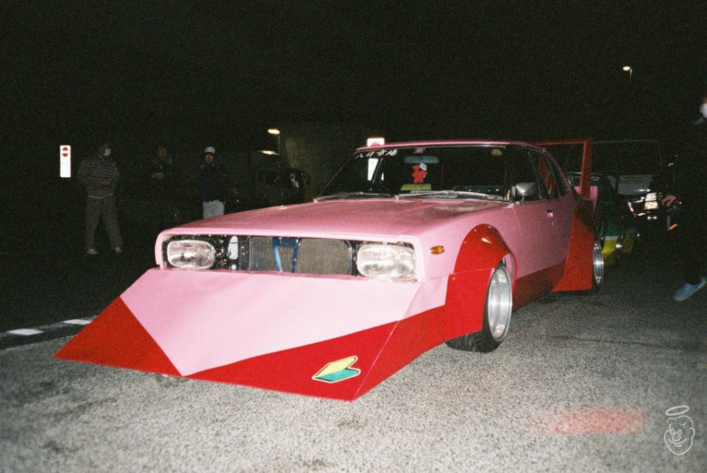 Custom pink and red Bosozoku car with Japanese text 八田ガレージ on the windshield at a nighttime gathering.