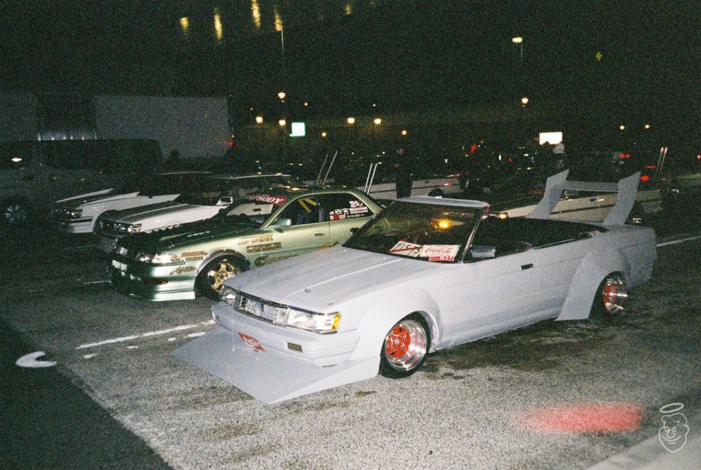 Modified Japanese drift cars parked at night with stickers displaying brand names like WORK and Coca-Cola on the windshields.
