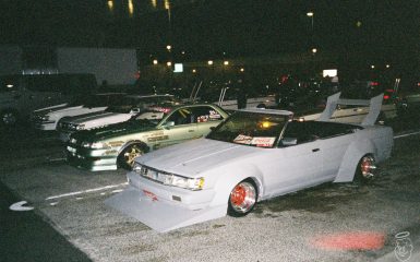 Modified Japanese drift cars parked at night with stickers displaying brand names like WORK and Coca-Cola on the windshields.