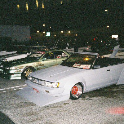 Modified Japanese drift cars parked at night with stickers displaying brand names like WORK and Coca-Cola on the windshields.