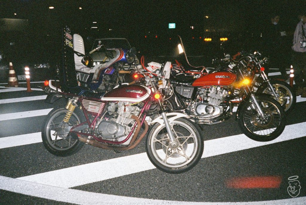 Three vintage motorcycles parked on a street at night, featuring vivid colors and custom modifications.