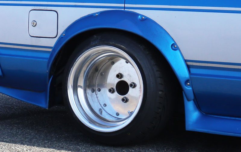 Close-up of a blue car with a wide rear wheel and a polished silver rim, showcasing custom fender flares.