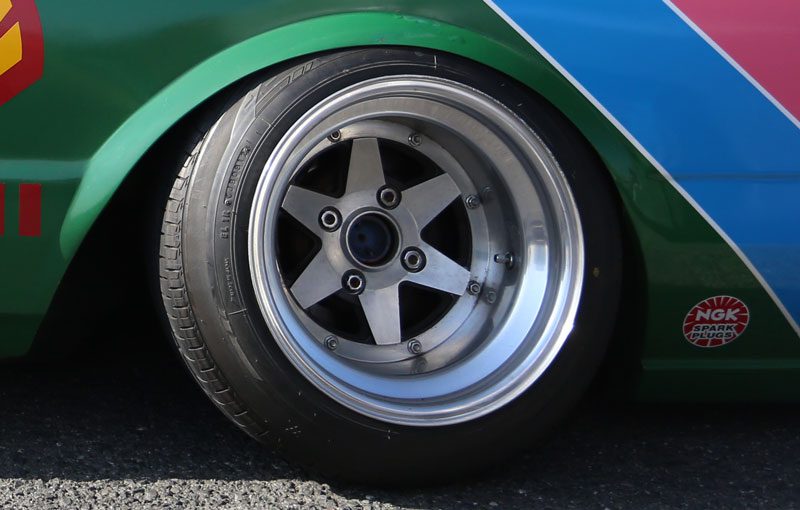 Close-up of a vintage car's alloy wheel and tire with NGK spark plugs sticker in a colorful backdrop.