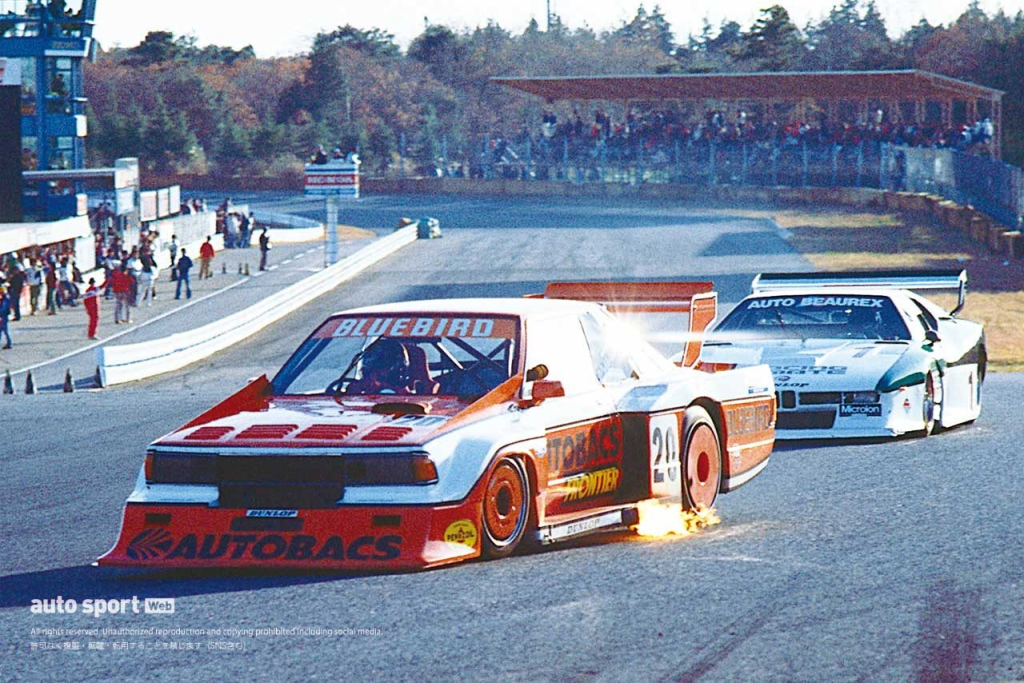 Race cars from AUTOBACS and AUTO BEAUREX speed down a track, with sparks flying from the lead vehicle's side.