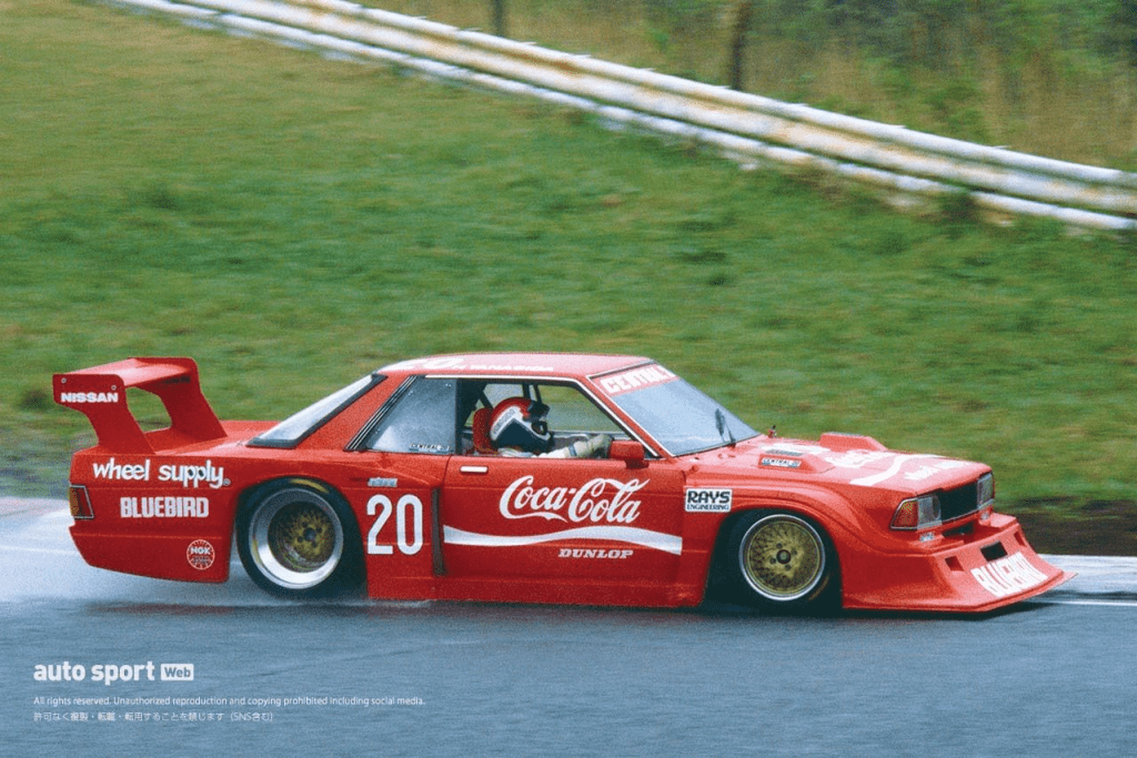 Red Nissan Bluebird race car with Coca-Cola branding, racing on a wet track. Car number 20 with wheel supply and DUNLOP.