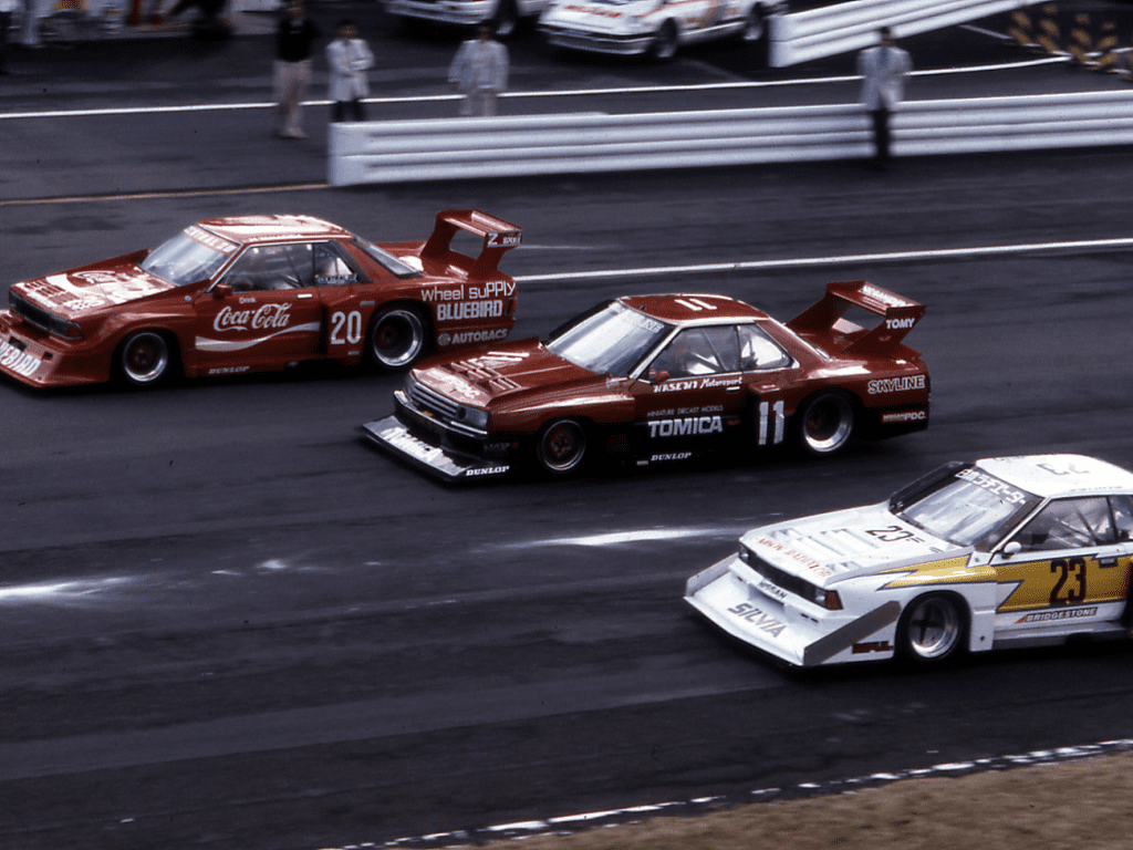 Three race cars competing on a track, featuring Coca-Cola, Bluebird, Tomica, and Silvia liveries with the numbers 20, 11, and 23.