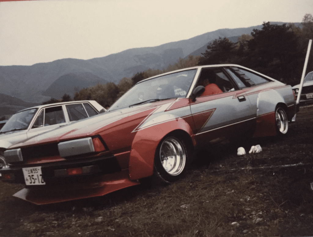 A modified red and silver Japanese car with wide wheels parked in a scenic mountain area, showcasing a vintage design.