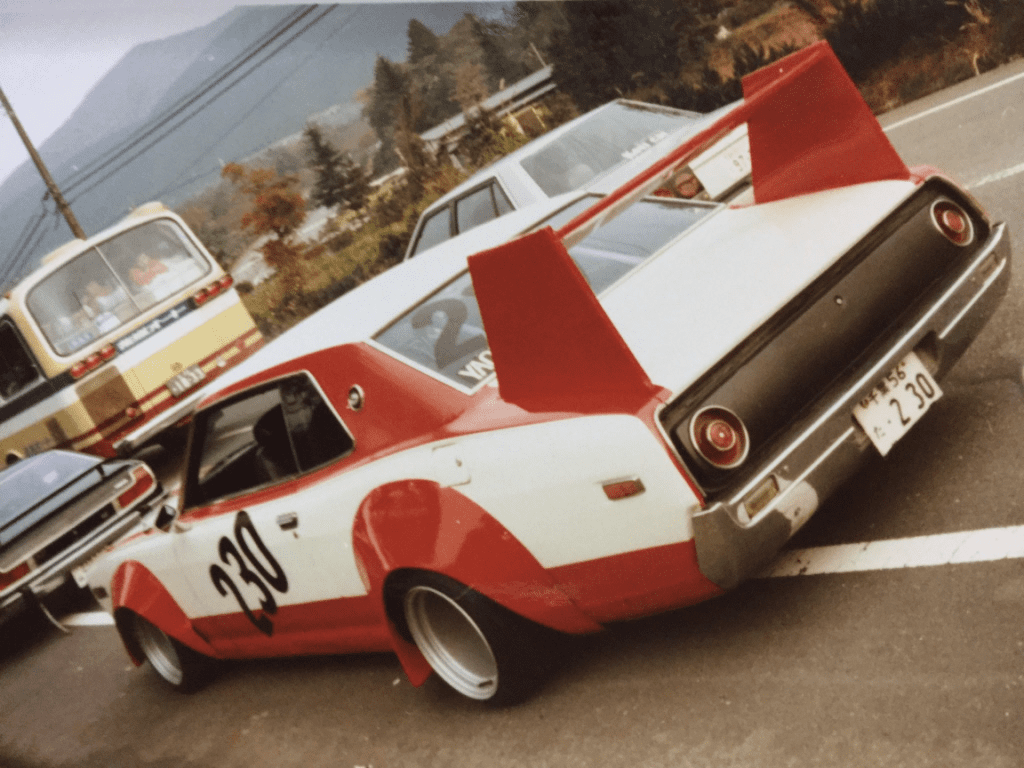 Vintage racing car with number 230 and bold red accents parked on a mountain road, surrounded by other vehicles.