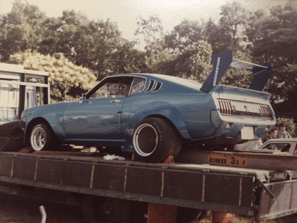 Vintage blue sports car with large spoiler on truck bed; Bridgestone brand on spoiler. Japanese text 多大重量車 3台本 for heavy vehicles.