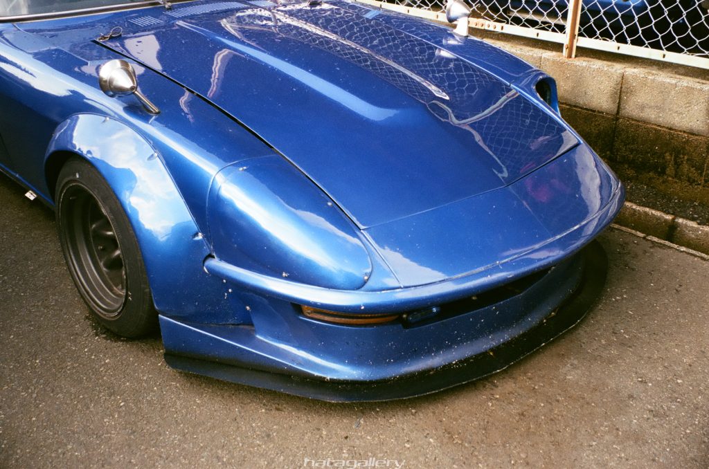 Blue classic sports car with custom wide fender flares and sleek front bumper parked on asphalt near a chain-link fence.