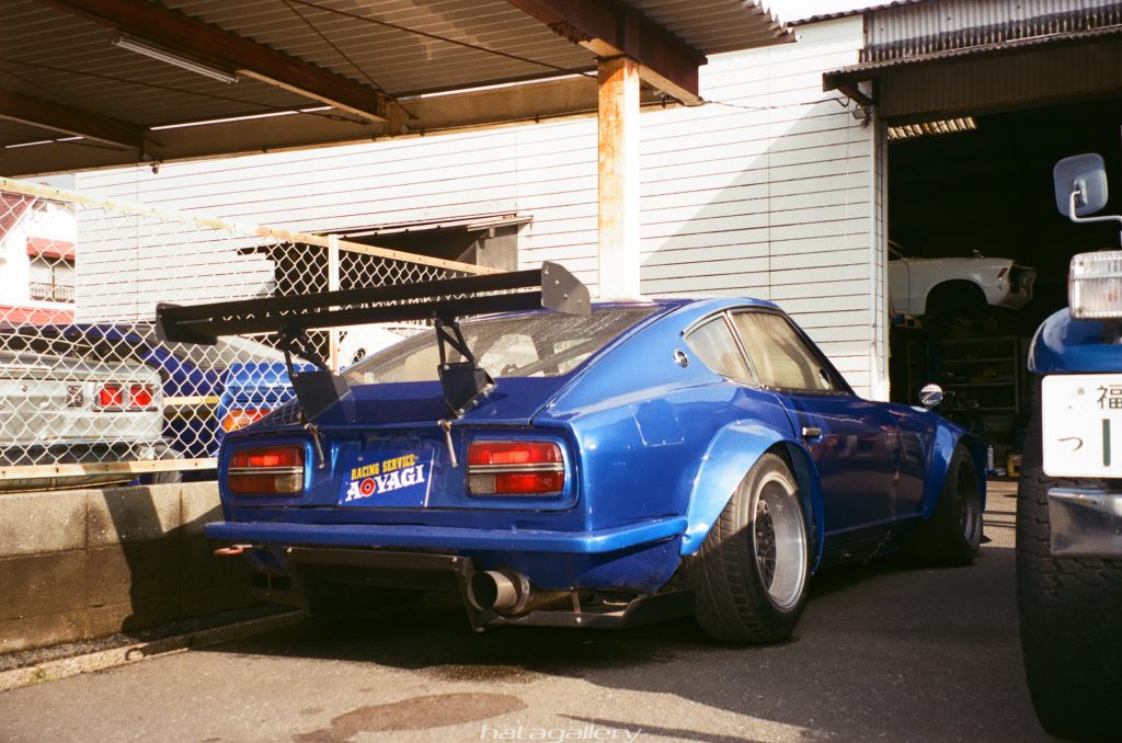 Modified blue retro sports car with Racing Service Aoyagi sticker, parked in Japanese garage lot.