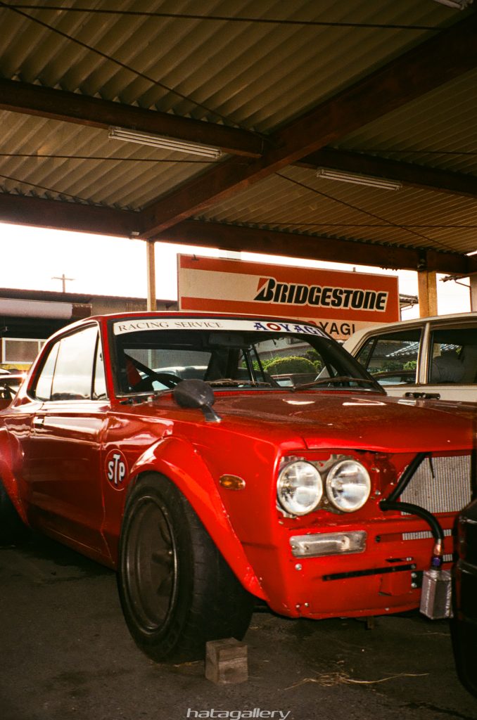 Red classic race car with Bridgestone sign in Japanese garage, featuring racing service AOYAGI and STP logo.