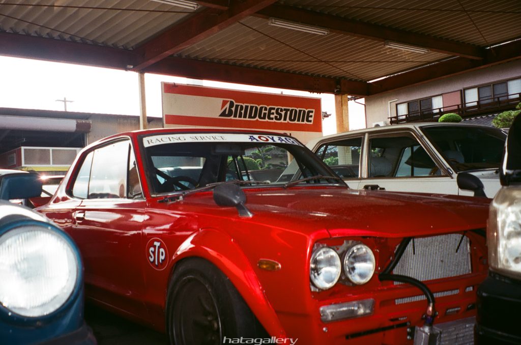 Red vintage racing car under a covered garage with Bridgestone sign, showing Racing Service and AOYAGI decals.