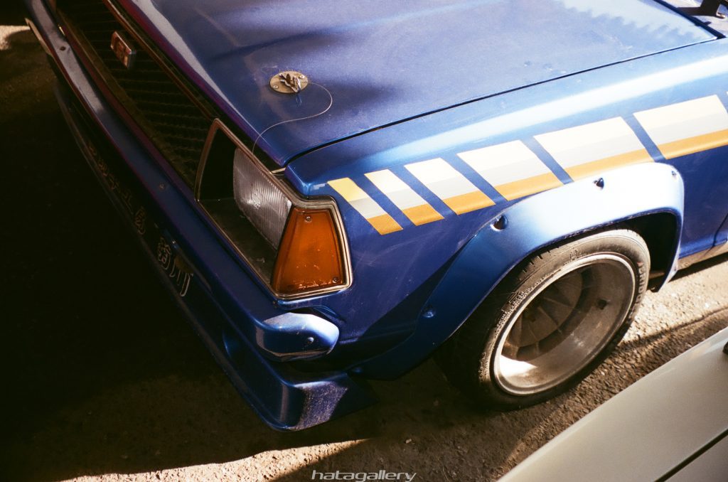 Close-up of a blue vintage car with yellow stripe detailing and Japanese text on the front bumper in a shaded area.